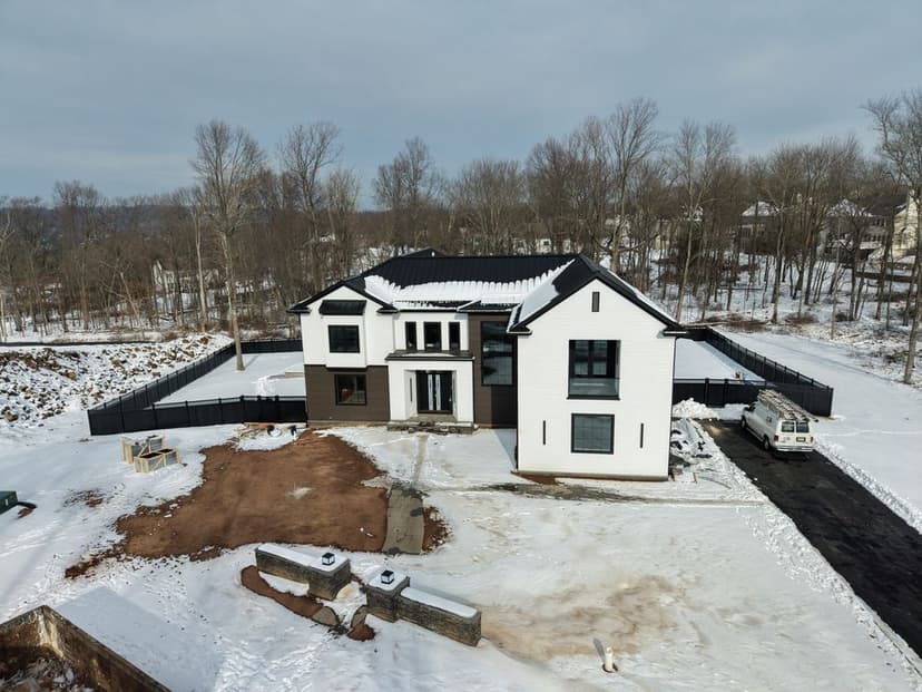 Modern two-story house under construction in winter landscape, surrounded by snow and bare trees.