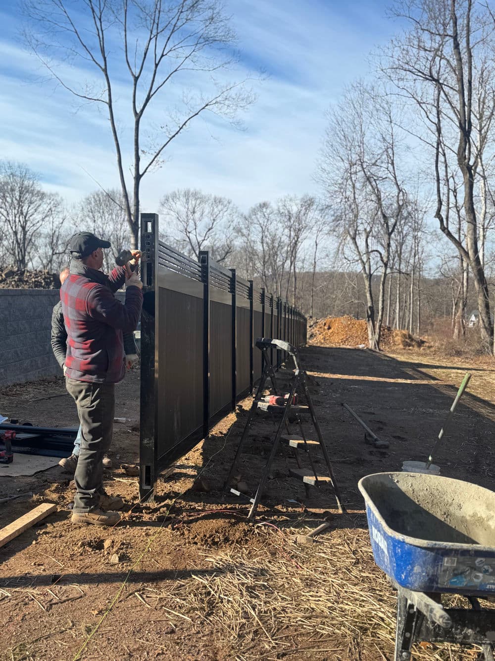 Man installing a modern fence in a rural area, surrounded by trees and construction tools.